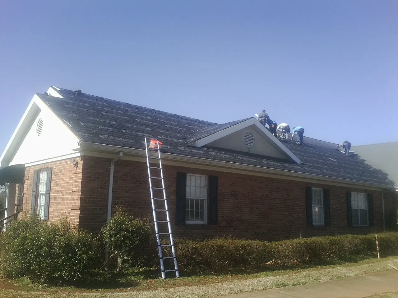 Crew tearing shingles off a brick ranch roof with a ladder set — Emergency Roof Repair work by our crew in Poneto