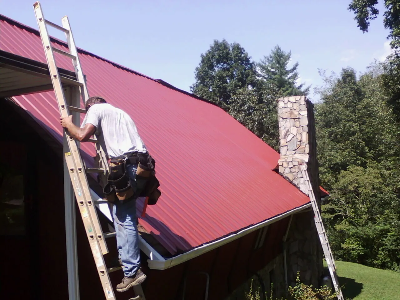 Roofer on a ladder beside a red metal roof and stone chimney — Soffit & Fascia Repair by Roofing Brothers in Poneto